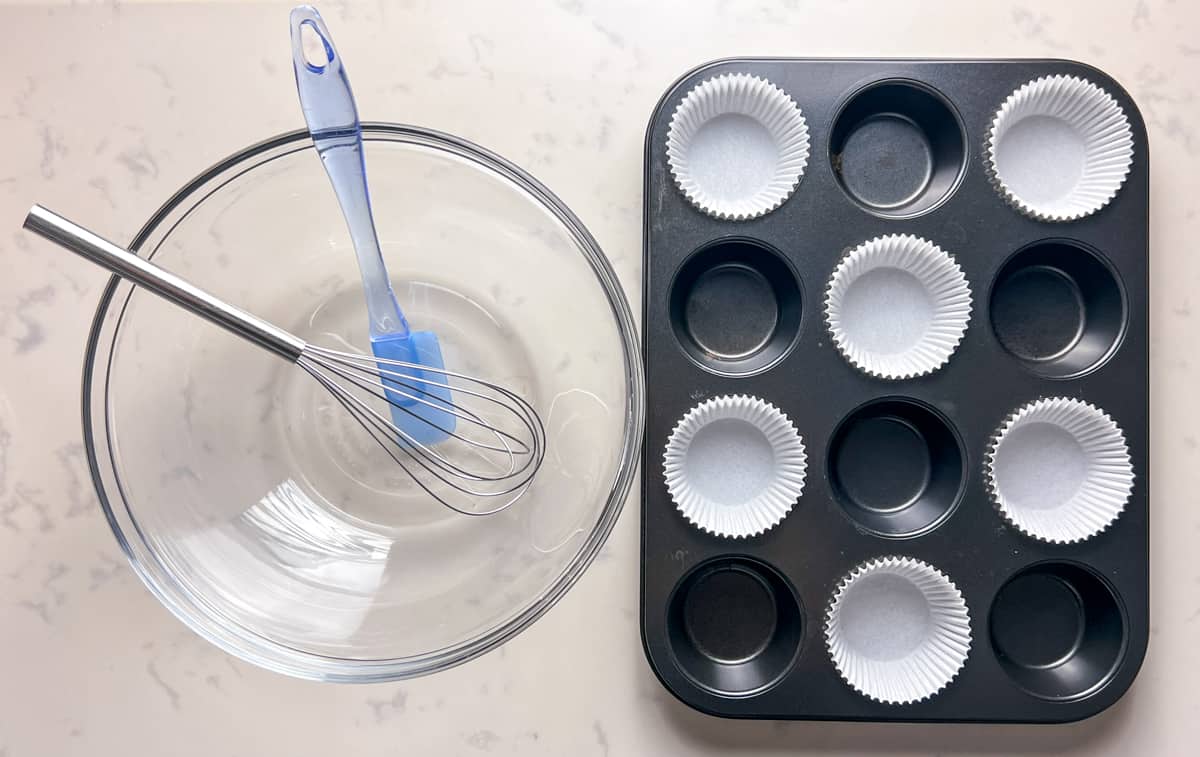 a bowl with whisk and spatula in it, next to a muffin pan with paper liners.