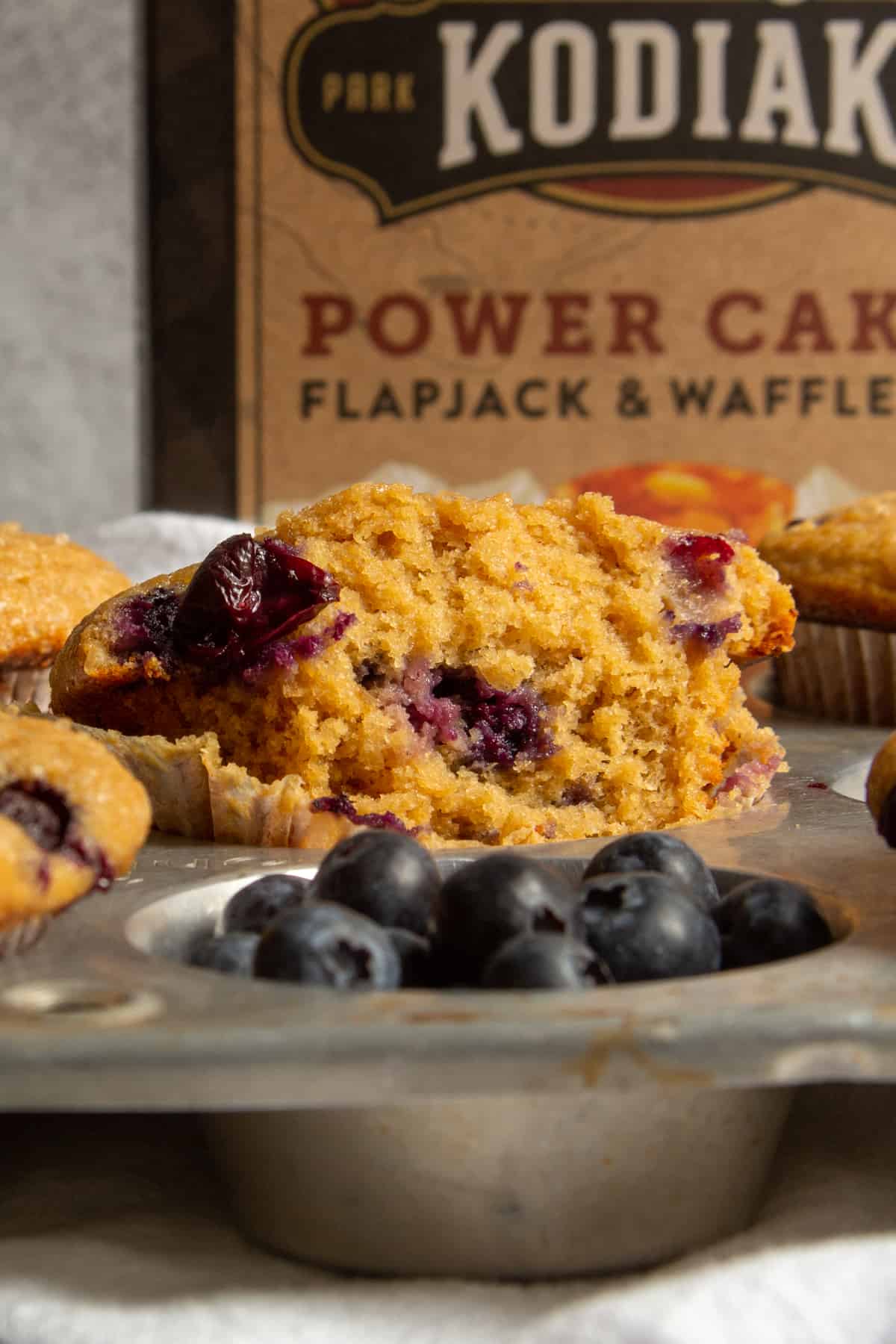 a blueberry muffin with paper wrapper removed sitting on a muffin pan and blueberries in front and the kodiak mix box in the back.