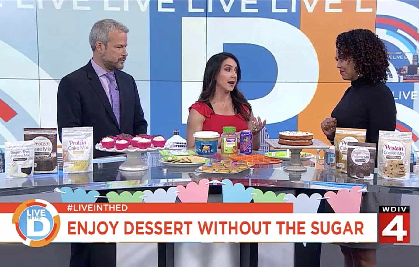 tv anchors behind a large table full of protein cakery baked treats.